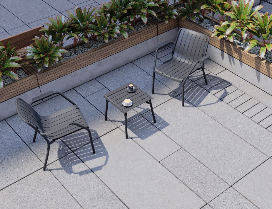 roku lounge sidetable charcoal An elevated view of an outdoor terrace with two dark-coloured chairs arranged around a small table. On one side, a long wooden planter box holds an arrangement of green plants with red-tinted leaves. The flooring is made of square grey tiles.