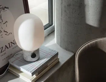 White marble JWDA Table Lamp resting on stacked books beside a window, framed artwork and a brown chair
