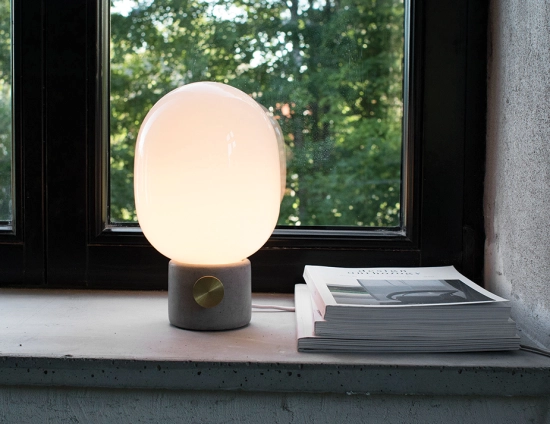 Glowing JWDA Concrete Table Lamp beside stacked books on a windowsill overlooking leafy trees