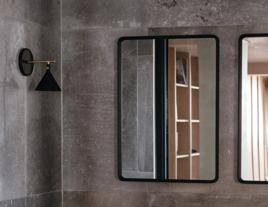 Black rectangular Norm Wall Mirrors on a grey tiled wall beside a black and gold wall lamp