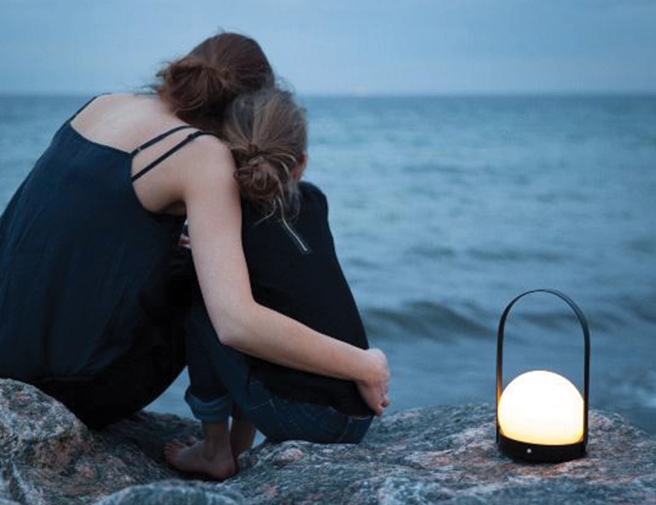 Glowing black Carrie Table Lamp beside two people sitting together on rocks by the sea