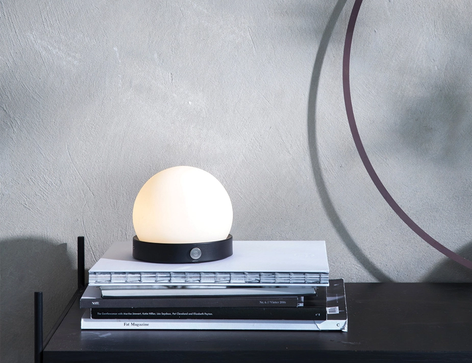 Glowing black Carrie Table Lamp with its handle removed, resting on stacked magazines beneath a round mirror